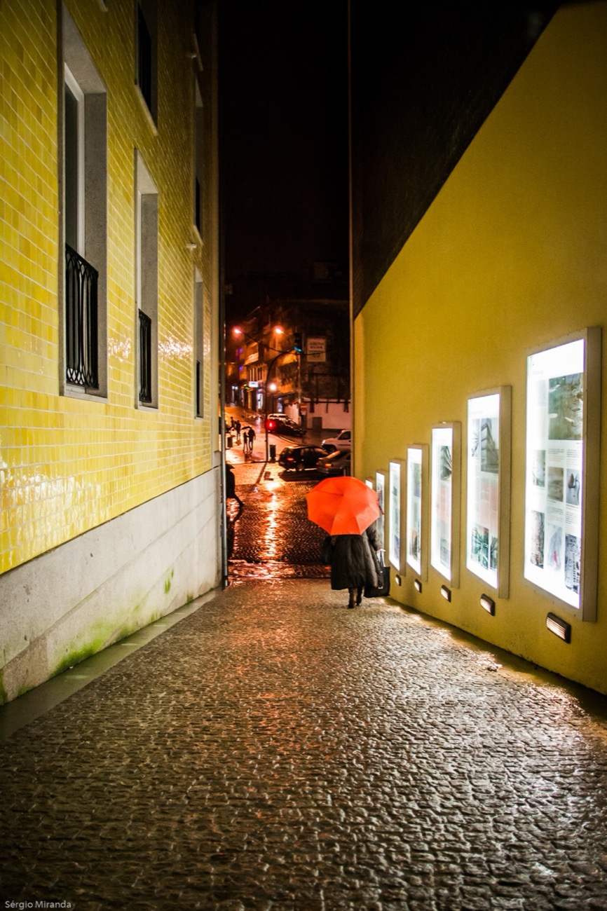 A narrow Porto street at night after rain, someone walking under a red umbrella